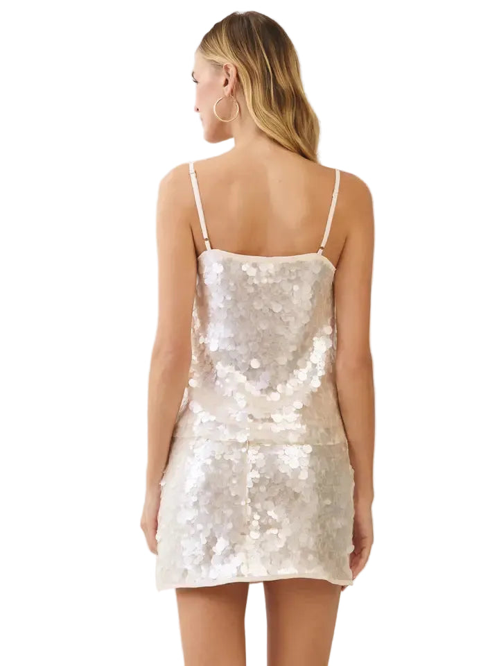 A woman shown from the back wearing a short, sleeveless sequined mini dress in a light champagne or off-white color, set against a plain light beige studio background.