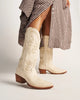 White cowboy boots with brown soles worn by a person in a checkered dress.