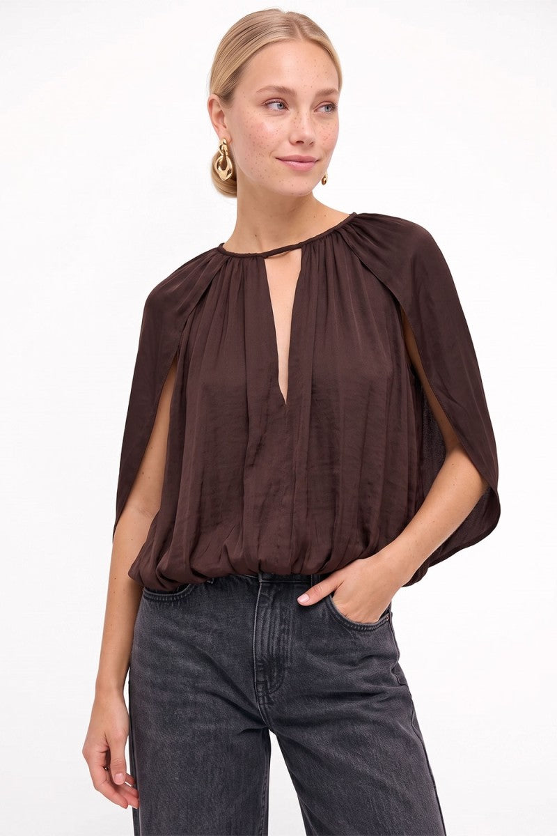 A woman wearing a dark brown draped blouse with a keyhole neckline and dark gray jeans, standing against a plain white background.