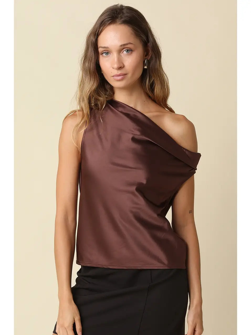 A woman modeling a dark brown satin one-shoulder top with a draped, asymmetrical neckline, paired with black pants, against a solid beige background.