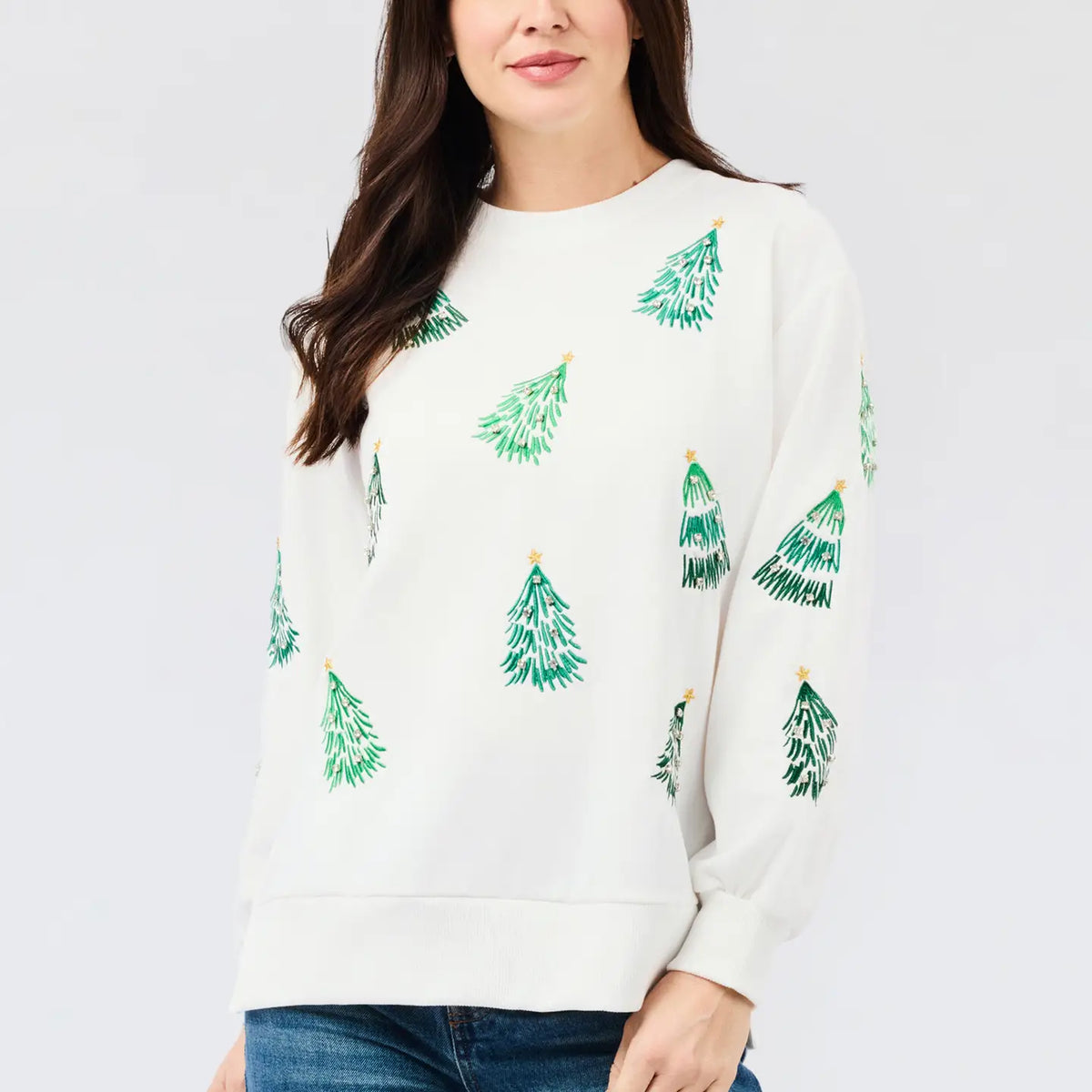 A woman wearing a cream-colored crewneck sweatshirt with a green Christmas tree pattern, paired with blue jeans, against a light gray studio background.