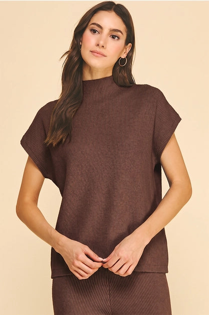 A woman modeling a ribbed knit two-piece lounge set in dark brown, featuring a short-sleeve mock neck top and matching pants, set against a solid beige background.