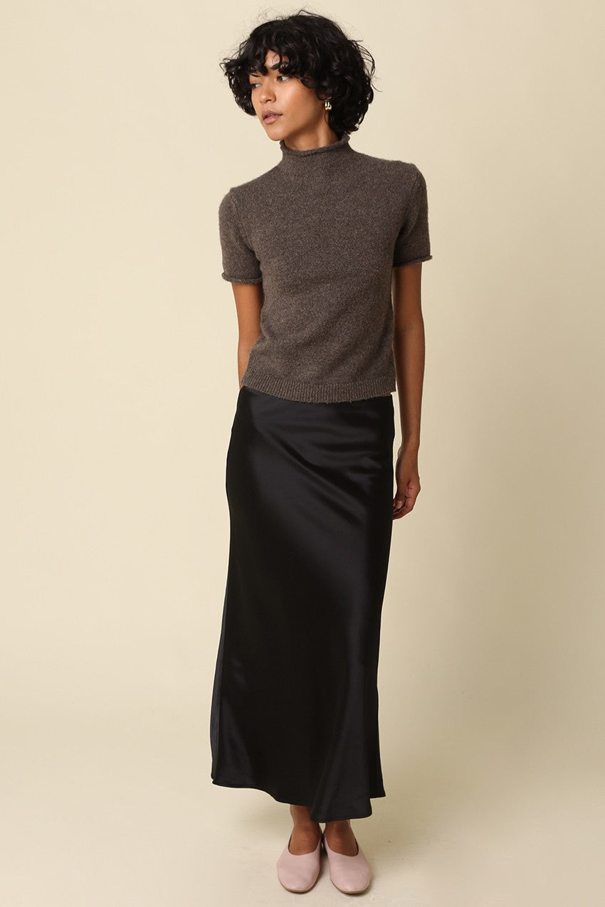 A model wearing a short-sleeve brown knit mock-neck top and a black satin midi skirt, paired with light pink flats, against a solid beige background.