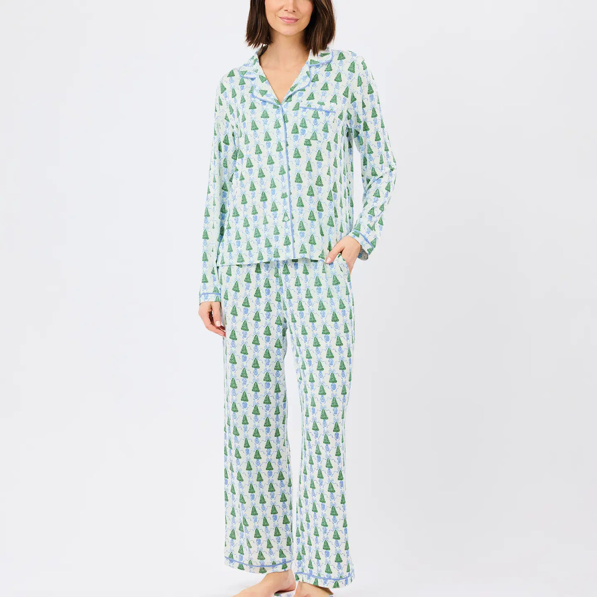 A woman modeling a matching pajama set with a light blue and green pine tree pattern, consisting of a long-sleeve button-up top and wide-leg pants, against a plain white studio background.