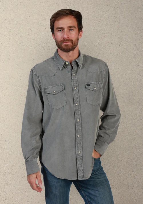 A man wearing a gray long-sleeve western-style button-down shirt with two chest pockets and blue jeans, standing against a textured beige wall.