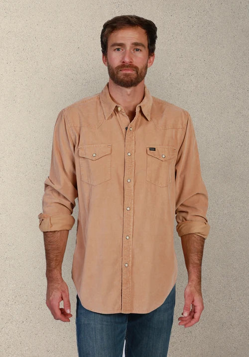 A man wearing a tan long-sleeve button-down shirt with two chest pockets and rolled-up sleeves, paired with blue jeans, standing against a textured light gray wall.