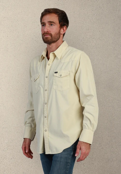 A man wearing a light cream-colored long-sleeve western-style button-down shirt with two chest pockets, paired with blue jeans, standing against a textured beige wall.