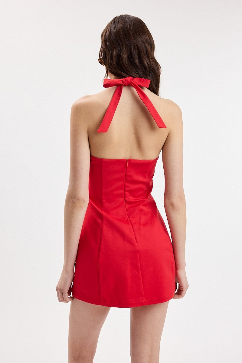 A woman shown from the back wearing a bright red halter mini dress with a structured back and bow detail at the neck, photographed against a plain light gray studio background.