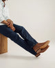 Person wearing dark blue jeans and brown shoes sitting on a wooden block against a plain background