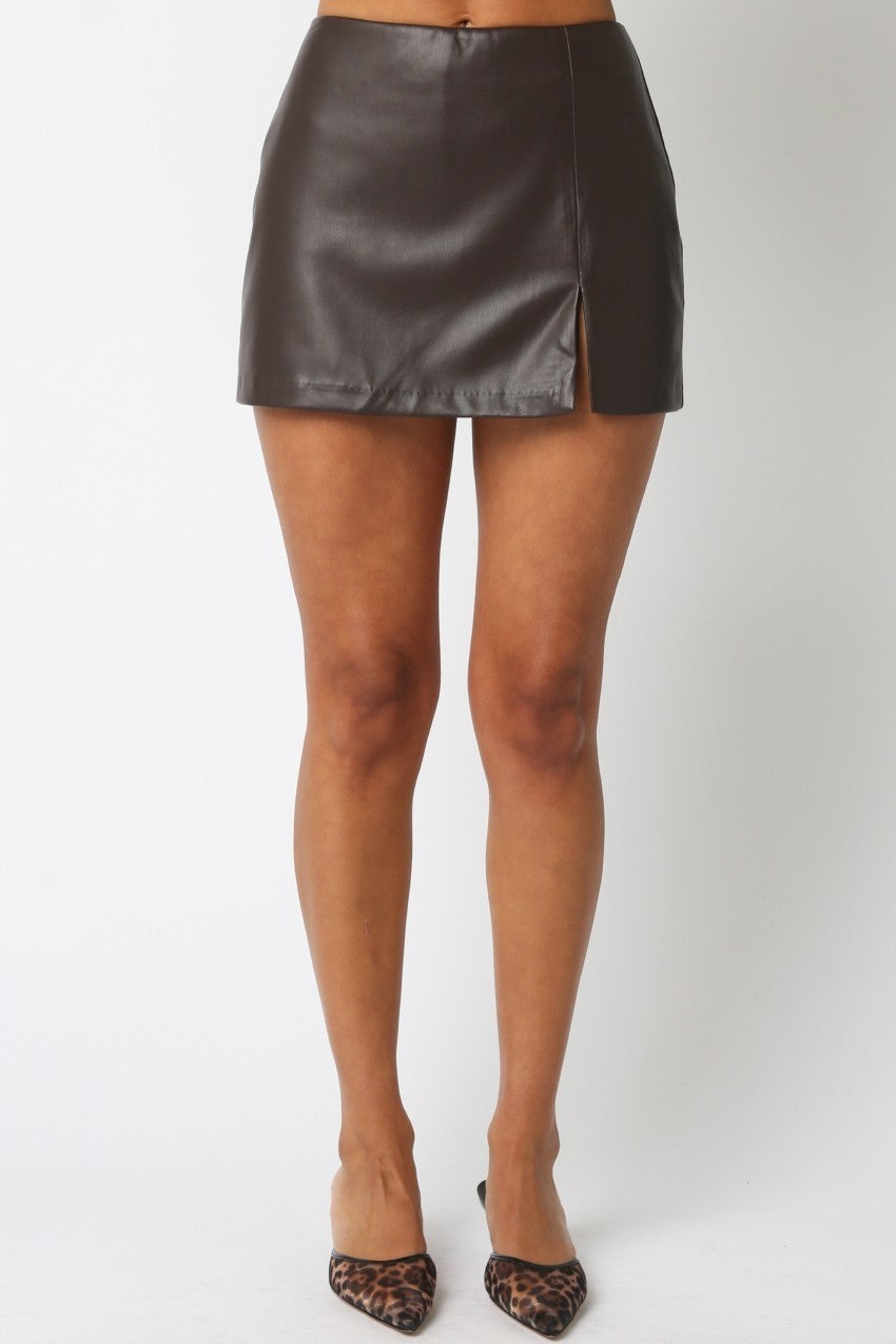A model wearing a dark brown faux-leather mini skirt with a side slit, paired with leopard print pointed-toe heels, shown against a plain white studio background.