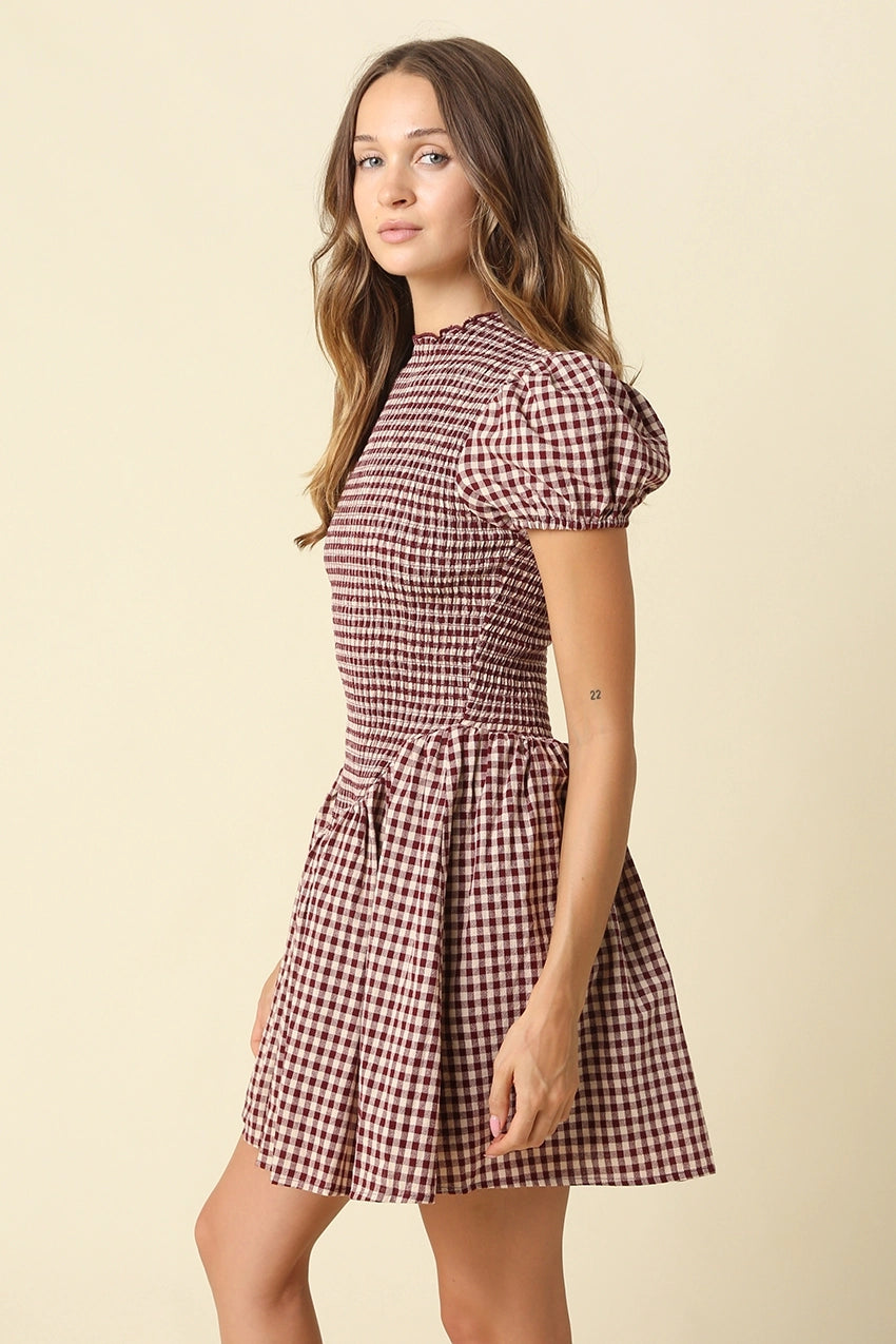 A woman modeling a short-sleeve mini dress with a red and beige gingham pattern, puff sleeves, and a gathered waist, set against a solid cream background.