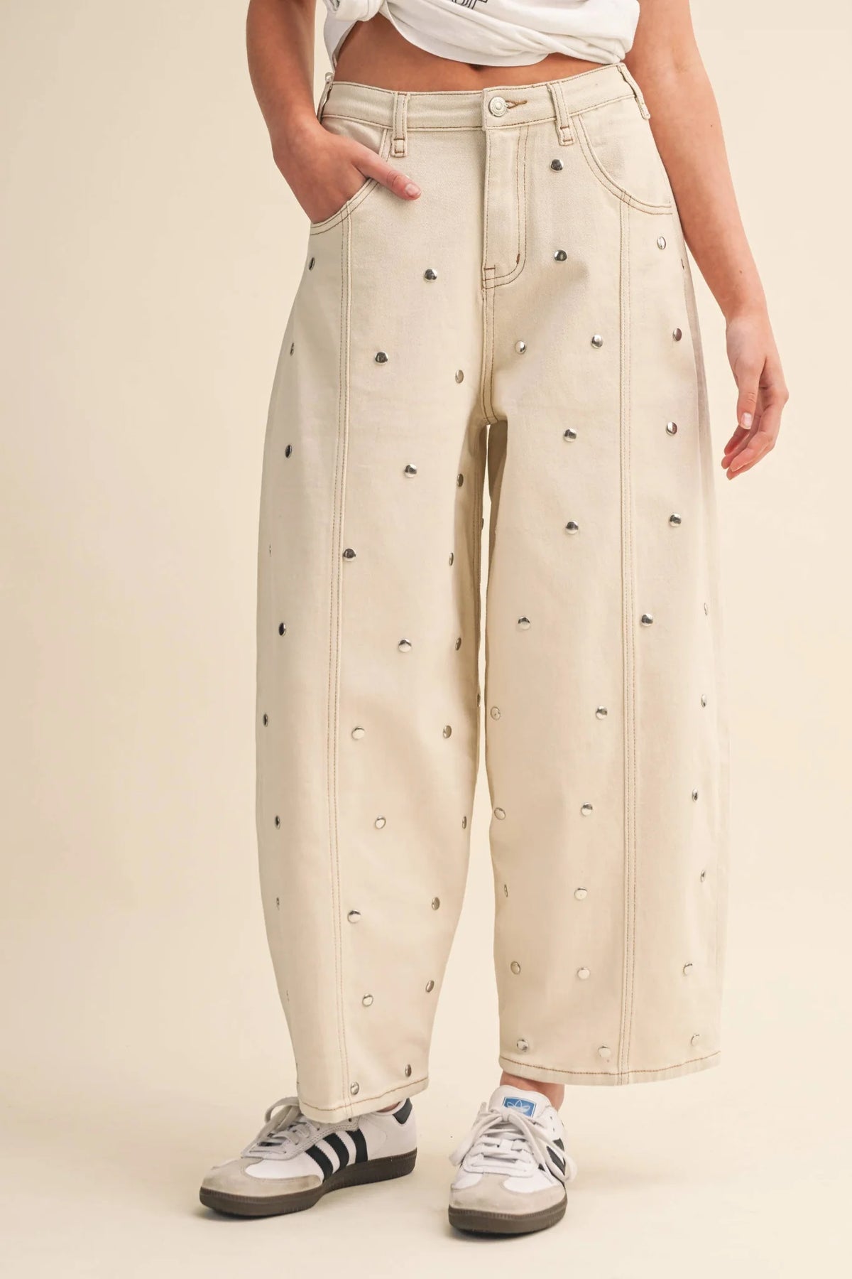 A model wearing high-waisted, wide-leg cropped jeans in a warm cream or off-white color, decorated with small black embroidered motifs and silver-tone studs. The model is also wearing a white cropped top and white sneakers with black stripes, set against a solid light beige studio background.