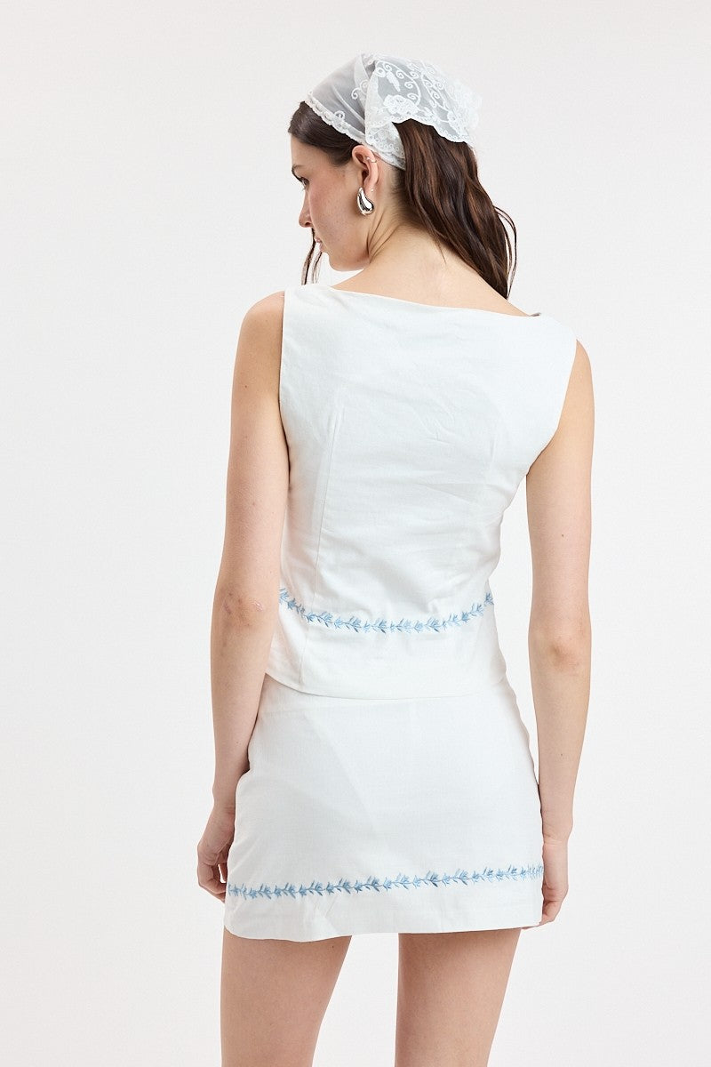 A woman shown from the back wearing a sleeveless white dress with light blue embroidered trim at the waist and hem, against a plain white studio background.