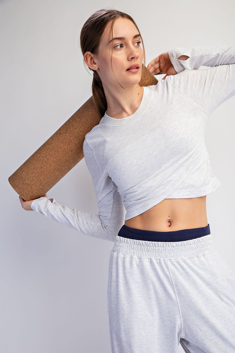 A woman modeling a light gray long-sleeve crop top and matching sweatpants, holding a rolled brown yoga mat over her shoulder against a plain light gray background.