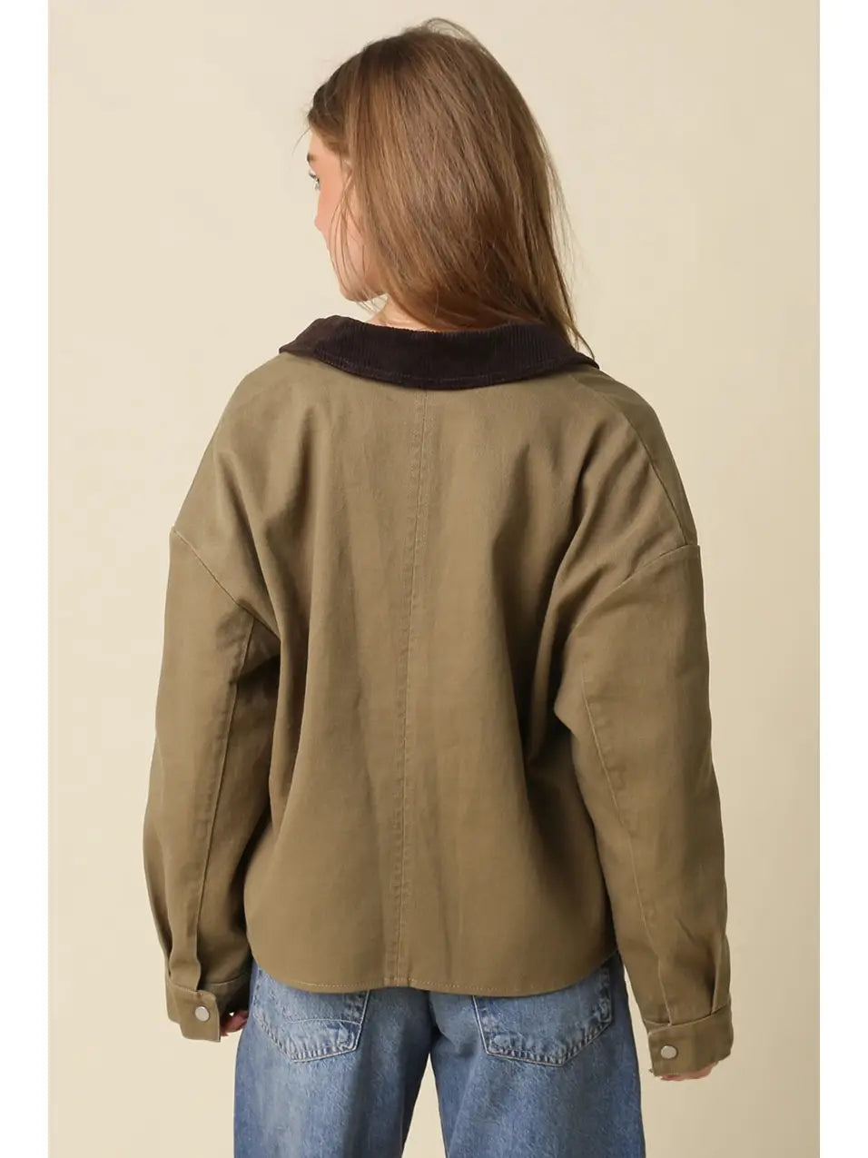 A woman viewed from the back wearing an oversized olive green jacket with a dark brown collar, paired with blue jeans, against a solid beige background.