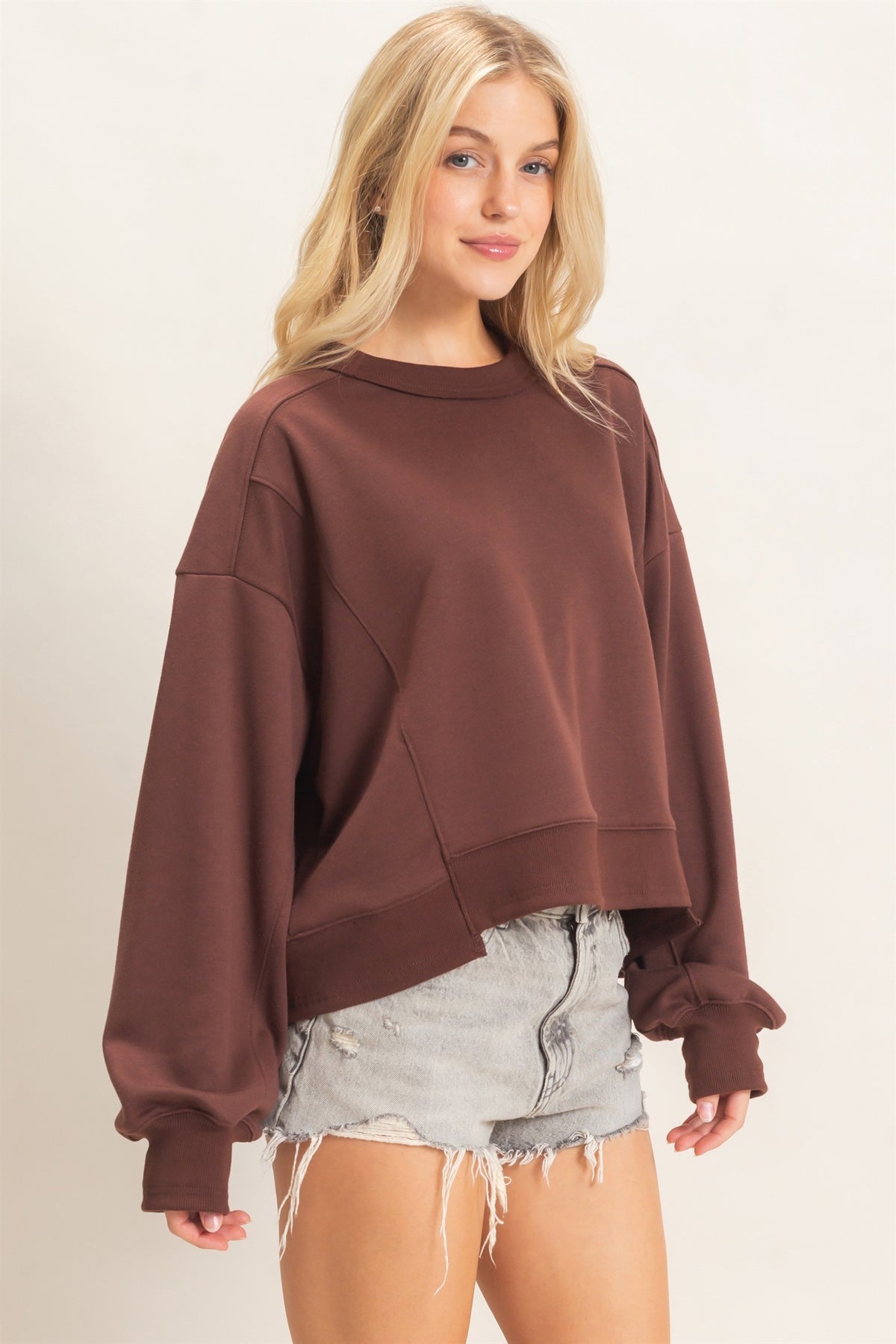 A woman modeling an oversized crewneck sweatshirt in a muted brownish-rose color, paired with frayed denim shorts, against a plain light background.