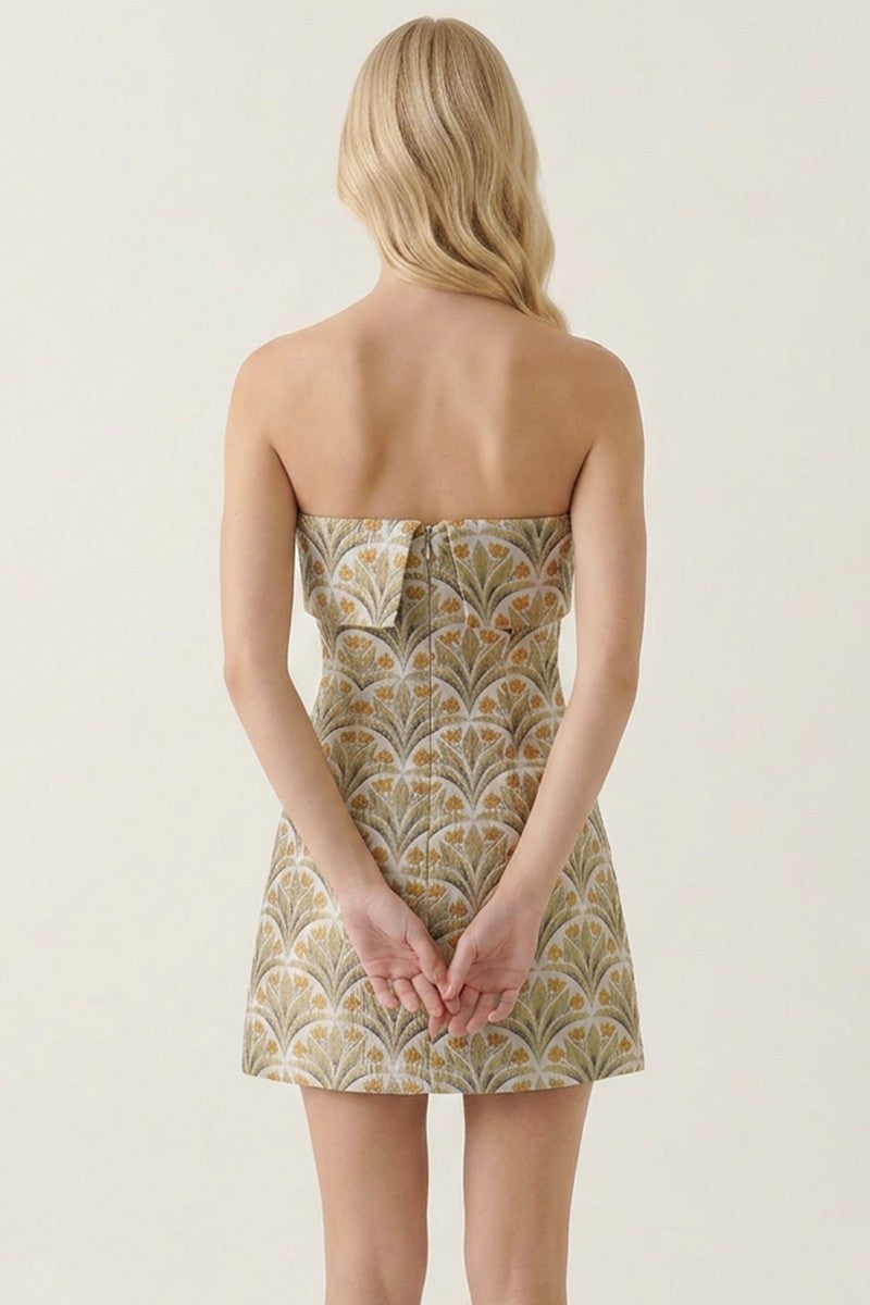 A woman shown from the back wearing a strapless mini dress with a beige, gold, and white Art Deco fan pattern against a plain light beige studio background.