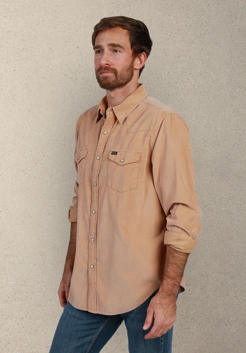 A man wearing a tan long-sleeve button-down shirt with two chest pockets and blue jeans, standing against a textured beige wall.