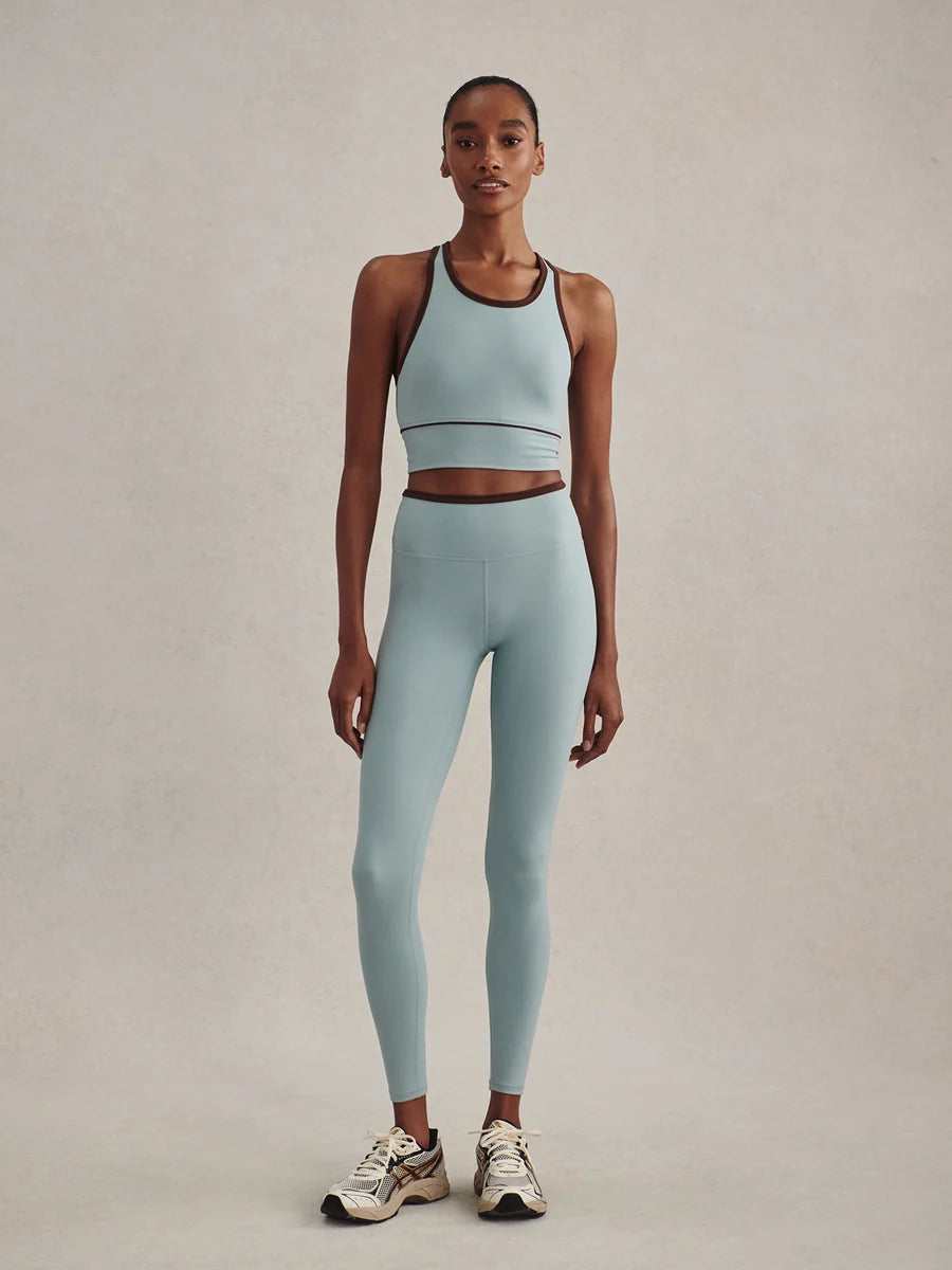 A model stands against a neutral beige studio background wearing a matching light blue athletic set consisting of a sports bra and high-waisted leggings, paired with white sneakers.