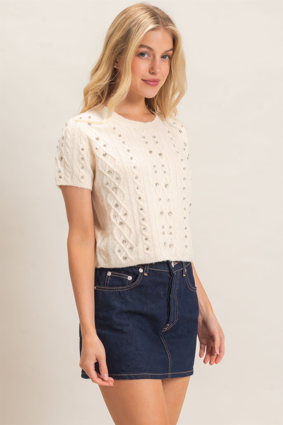 A woman modeling a cream-colored short-sleeve knit top with textured cable and eyelet patterns, paired with a dark blue denim mini skirt, against a plain light beige background.