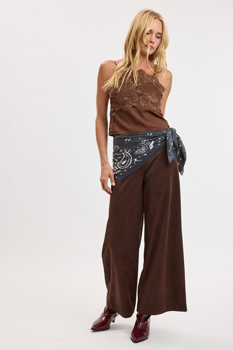 A woman modeling a brown sleeveless top with a lace overlay, wide-leg brown pants, a dark gray patterned sash tied at the waist, and dark brown pointed-toe boots, photographed against a plain white studio background.