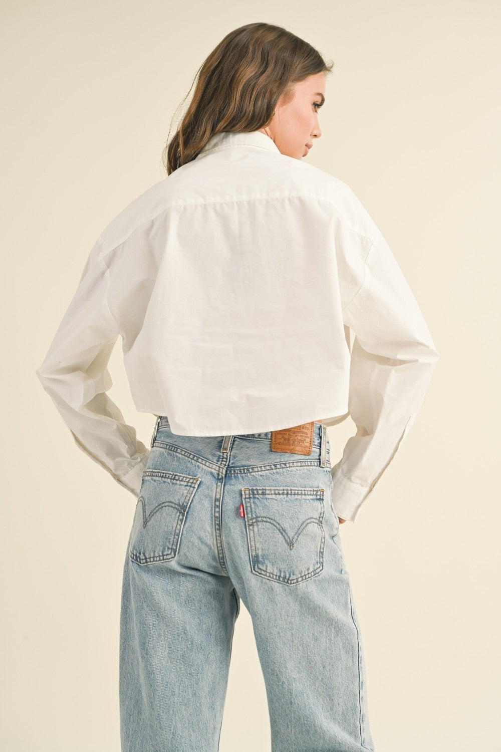 Woman standing on tan background wearing a cropped white button front shirt and jeans