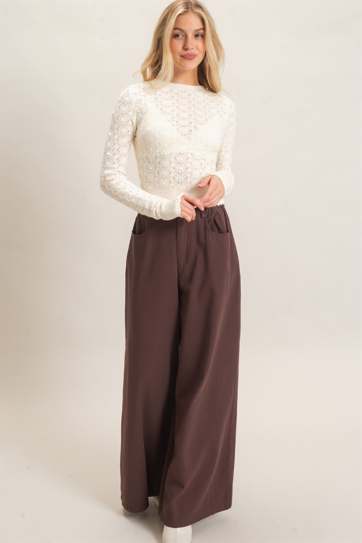 A woman modeling a cream-colored long-sleeve lace top paired with high-waisted, wide-leg brown trousers, set against a plain light beige studio background.