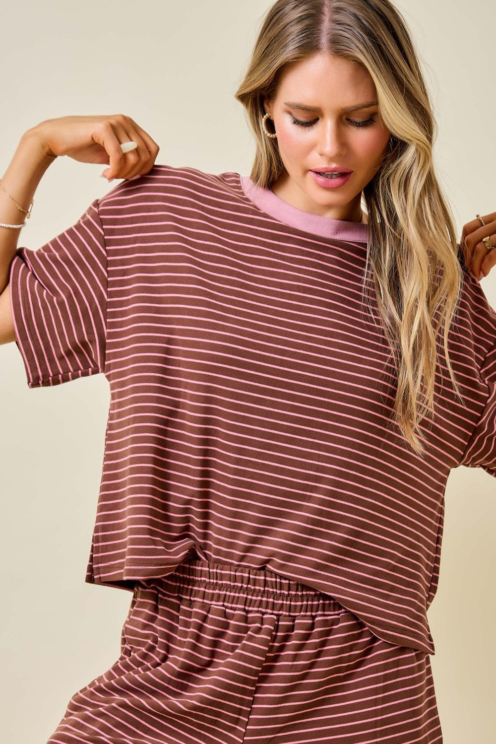 A woman wearing a loose short-sleeve top and matching pants set in a dusty rose color with thin white horizontal stripes, against a solid beige background.