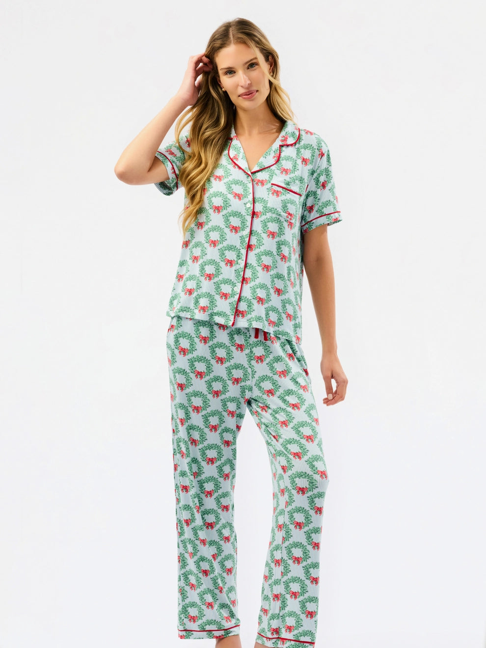 A woman modeling a two-piece pajama set with a green and white holiday wreath pattern and red trim, set against a plain white studio background.