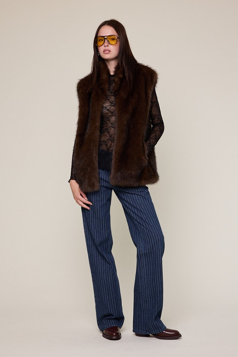 A model stands against a plain beige studio background wearing a dark brown fur vest over a black lace top, navy pinstriped trousers, and brown leather shoes.