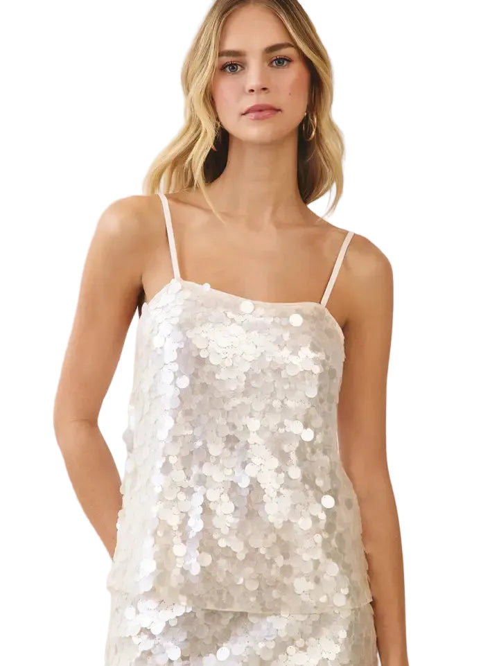 A woman modeling a sleeveless sequined camisole top in a light champagne or off-white color, featuring thin spaghetti straps and a tiered ruffle hem, set against a plain light beige background.