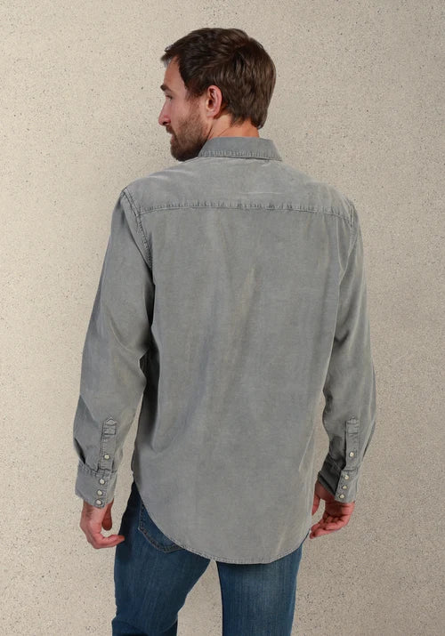 A man viewed from behind wearing a light gray long-sleeve button-up shirt and blue jeans, standing against a textured beige wall.