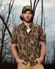 Man standing in front of trees wearing a camo short sleeve button down,khakis, and a camo hat