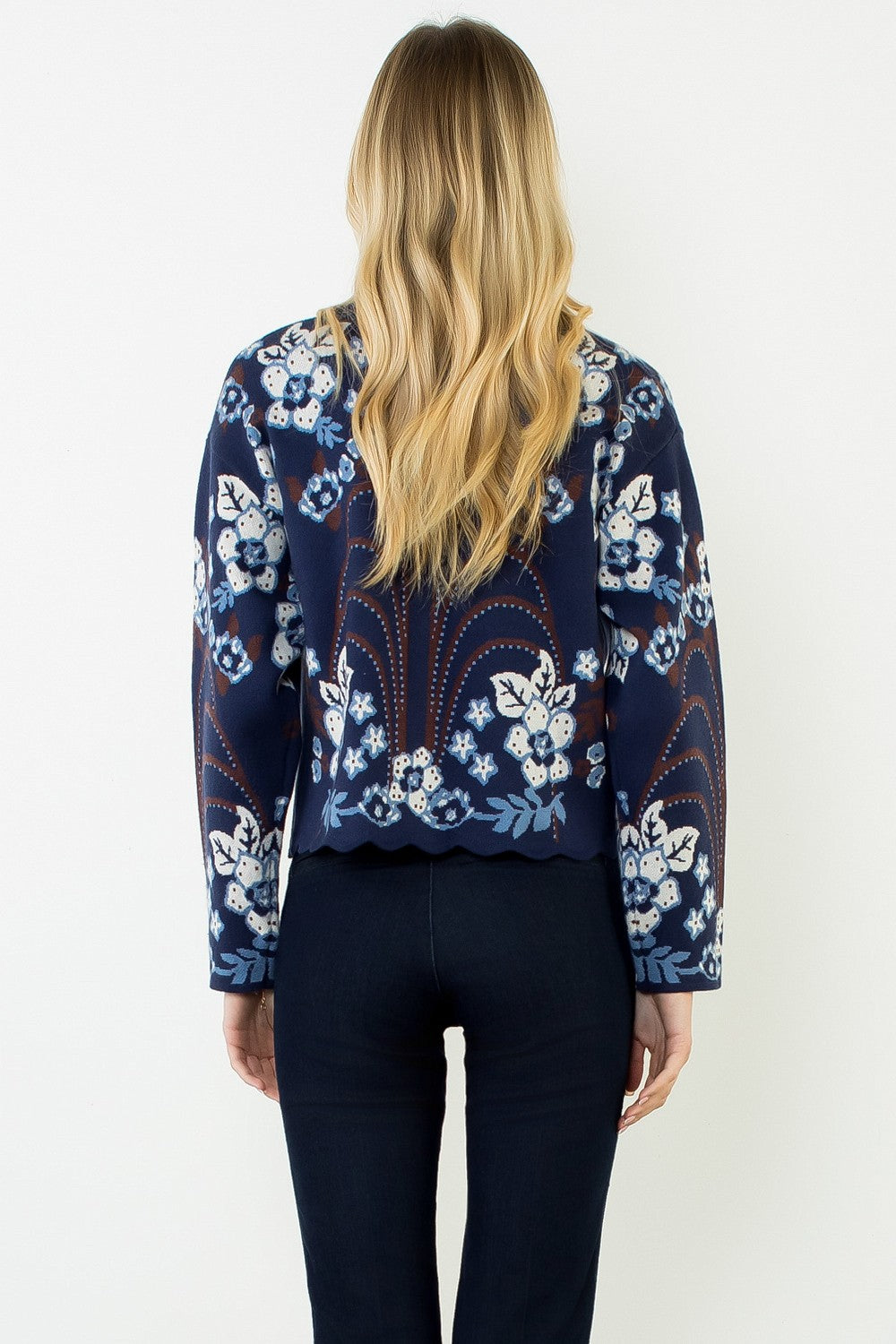 blonde woman standing in dark jeans modeling a navy sweater with floral pattern across the whole thing including sleeves. with a scallop hem