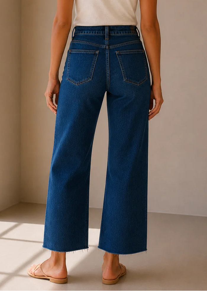 A woman viewed from behind wearing dark blue wide-leg cropped jeans, a white t-shirt, and tan flat sandals in a minimalist beige room with soft window light.