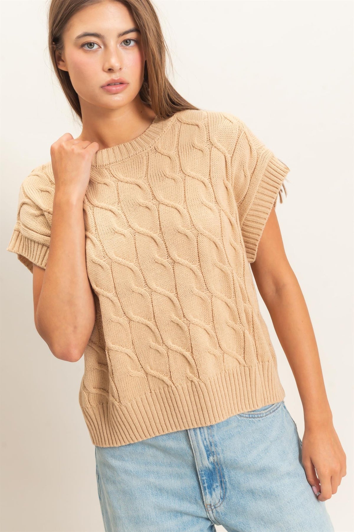 A woman wearing a beige cable-knit short-sleeve sweater and light blue jeans, photographed against a plain light background.
