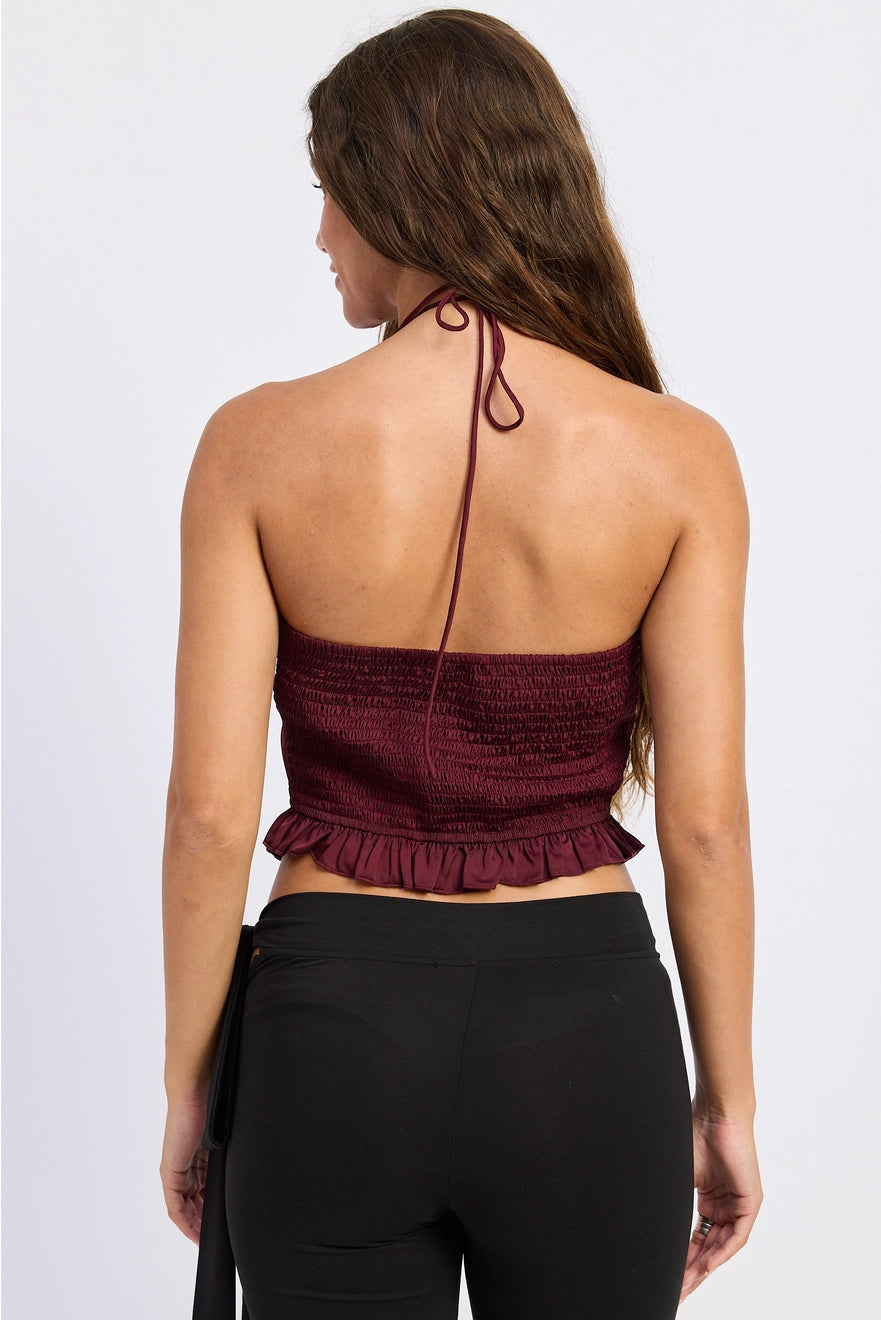A woman shown from the back wearing a dark burgundy halter crop top with a smocked texture and ruffled hem, paired with black leggings, against a light gray studio background.