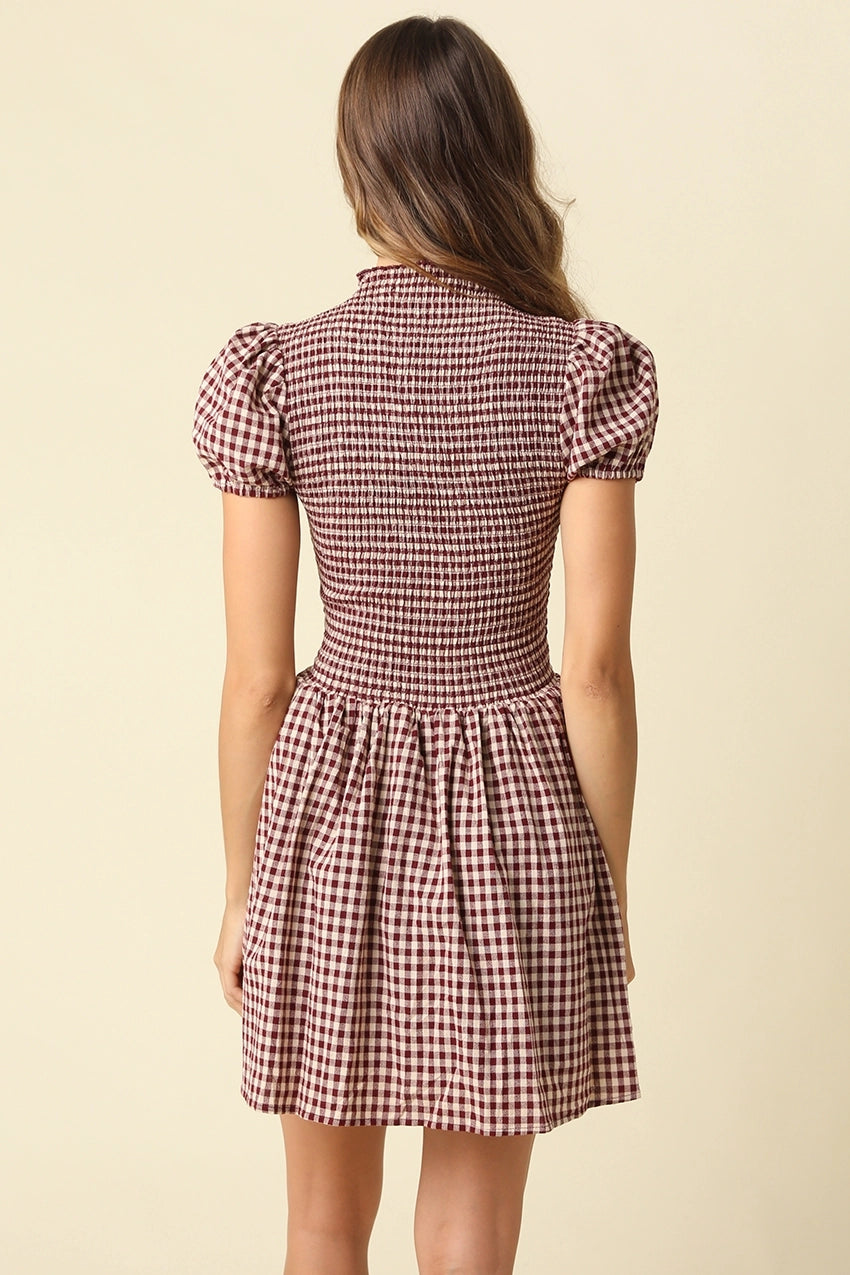 A woman viewed from the back wearing a short puff-sleeve dress with a brown and beige gingham pattern against a solid cream background.