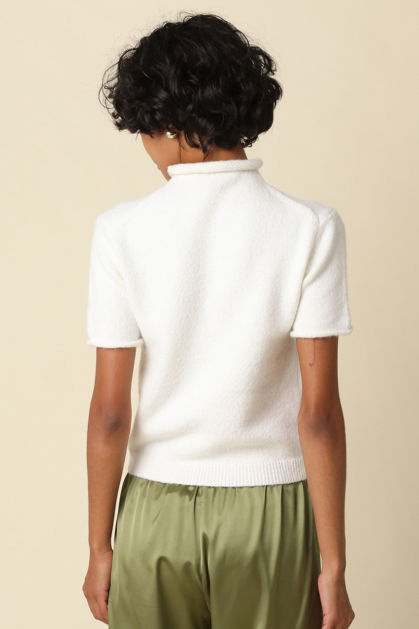 A person shown from the back wearing a short-sleeve white knit mock-neck or turtleneck top with olive green satin-like pants, against a plain warm beige studio background.