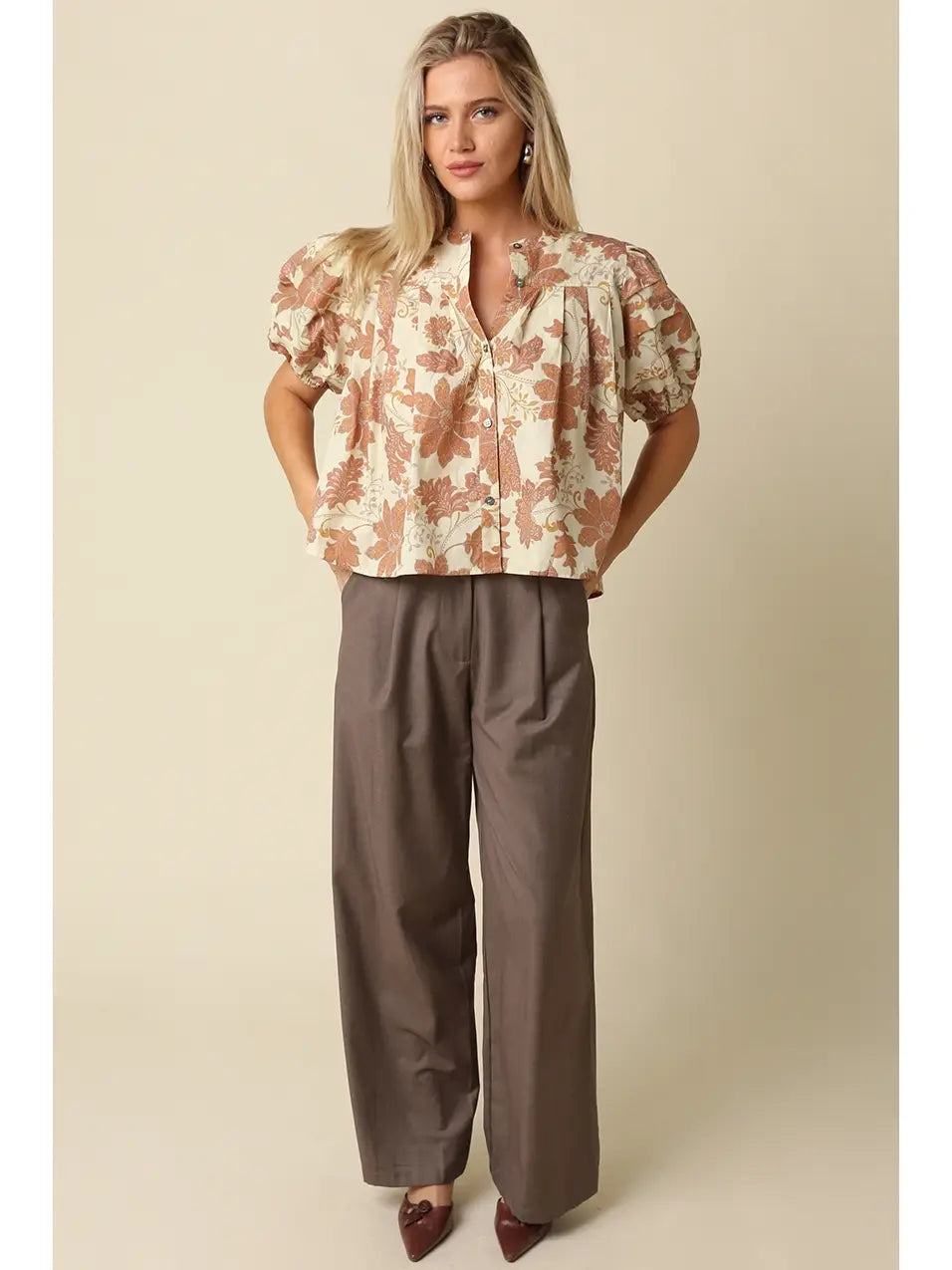 A woman modeling a short-sleeve button-up blouse with a beige and brown floral pattern, paired with wide-leg taupe pants, against a solid beige studio background.