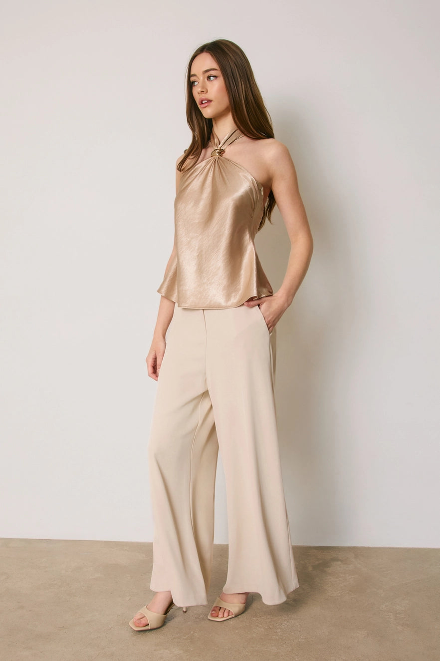 A woman modeling a sleeveless metallic champagne halter top paired with wide-leg cream pants and nude flat sandals, standing against a neutral light gray wall.