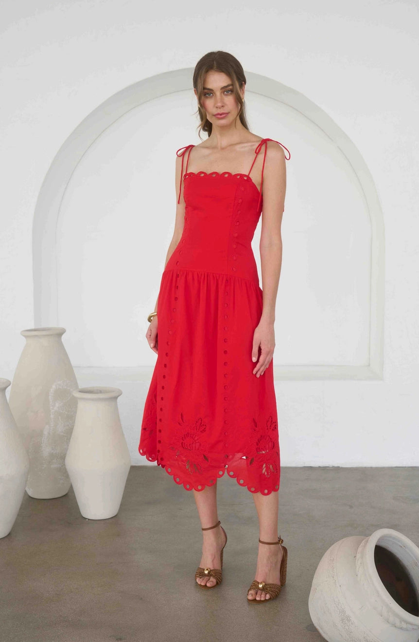 A woman modeling a bright red midi dress with thin shoulder straps, scalloped edges, and embroidered details, standing in front of a white arched wall with decorative ceramic vases.