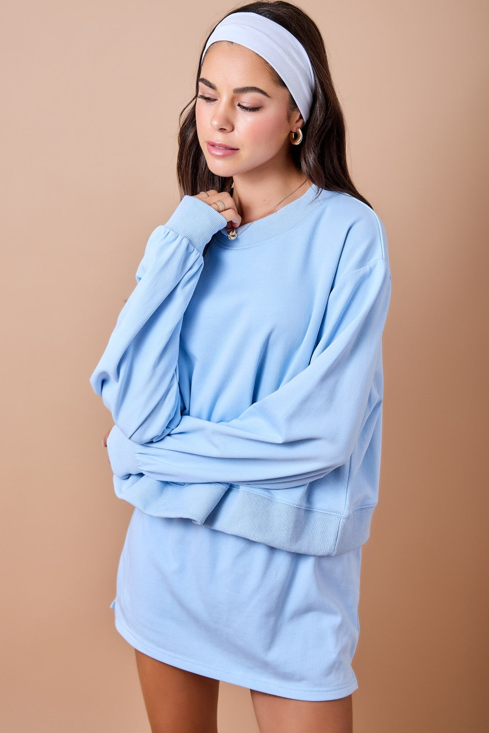 A woman wearing an oversized light blue sweatshirt and matching shorts set, styled with a white headband, against a solid beige studio background.