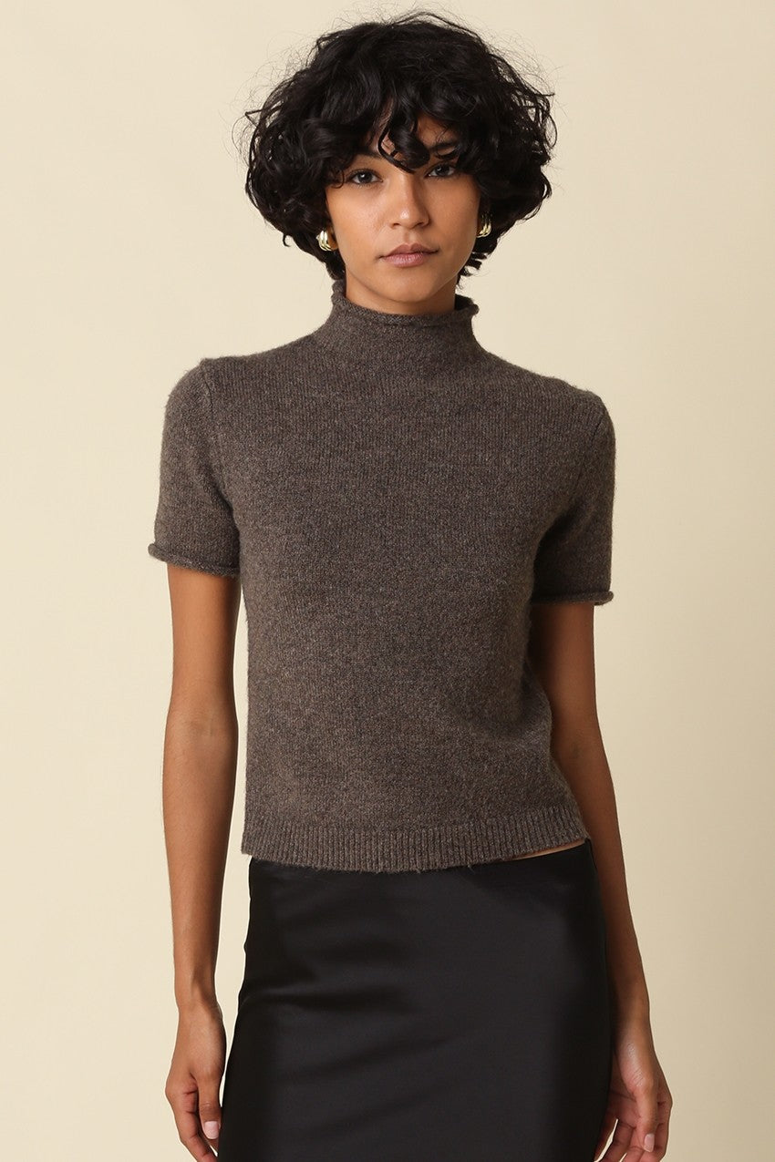 A woman wearing a short-sleeve ribbed mock neck knit top in a dark brownish-gray color, paired with a black skirt, against a solid beige background.