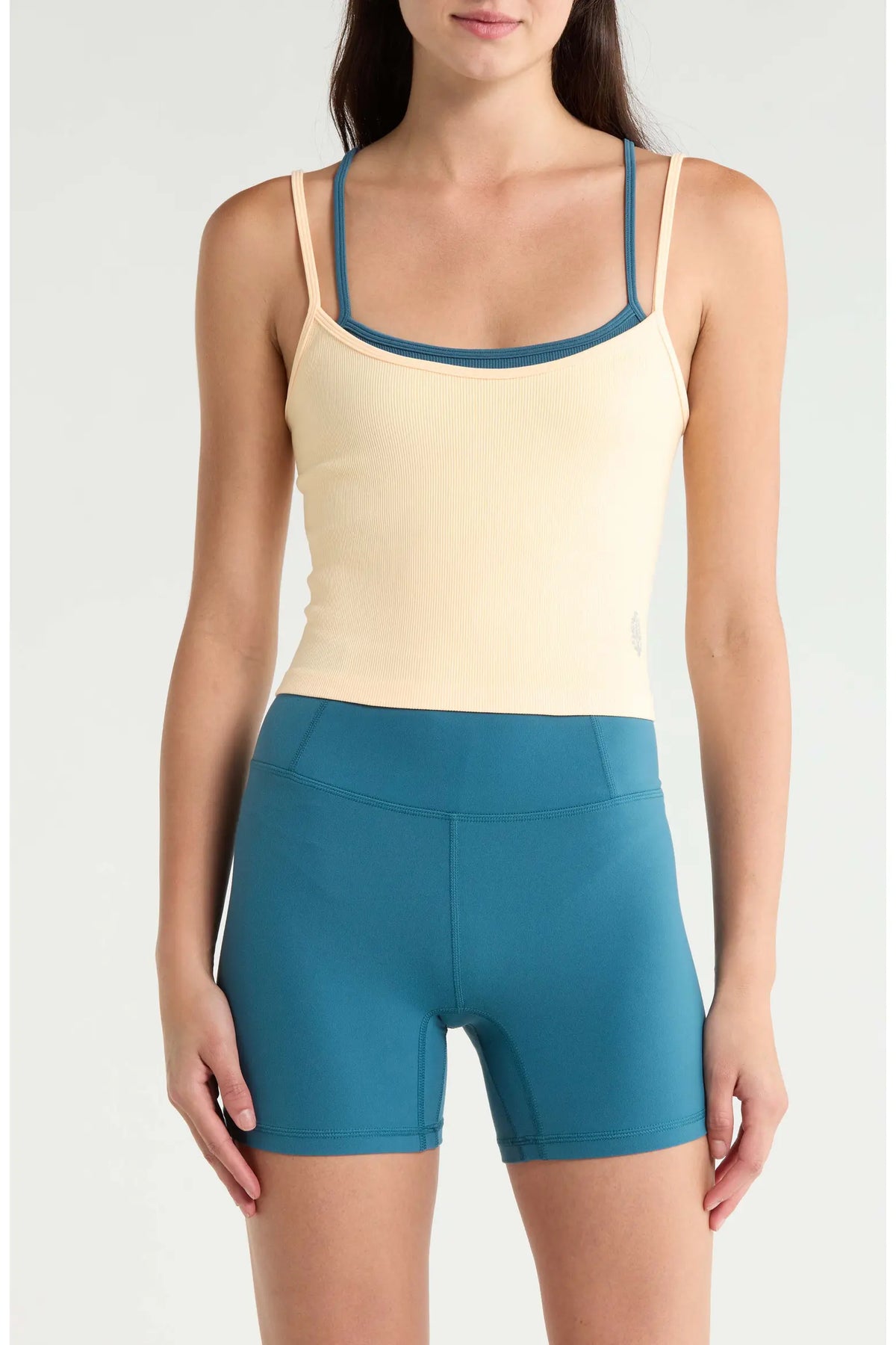 A model wearing a light cream ribbed camisole tank top with teal trim and matching teal high-waisted athletic shorts, shown against a plain light gray studio background.