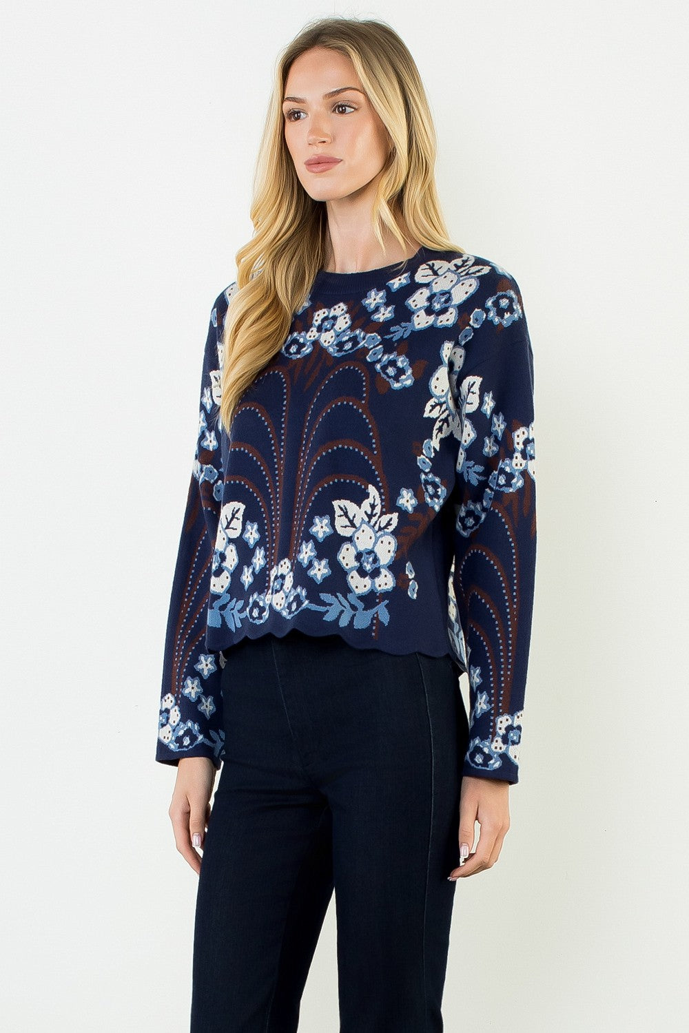 blonde woman standing in dark jeans modeling a navy sweater with floral pattern across the whole thing including sleeves. with a scallop hem