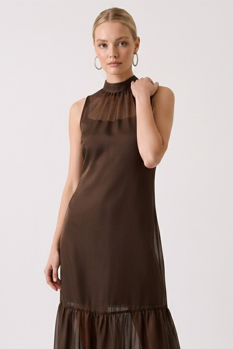 A woman modeling a sleeveless high-neck brown dress with sheer mesh panels, photographed against a plain light gray studio background.