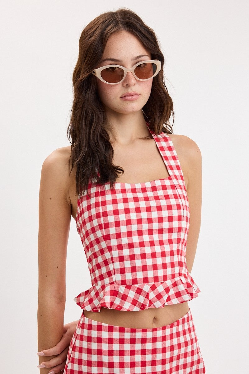 A woman modeling a red and white gingham check two-piece set consisting of a halter crop top with a ruffled hem and matching high-waisted bottoms, paired with cream cat-eye sunglasses, against a plain white background.