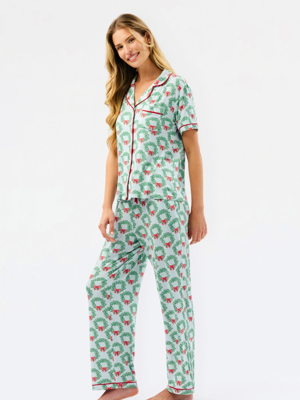 A woman modeling a two-piece pajama set with a green wreath pattern and red trim, featuring a short-sleeve button-up top and matching pants, against a plain white background.