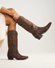 Woman pulling brown boots on in front of a white backdrop. 